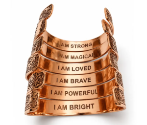 Stos miedzianych bransoletek ułożonych tak, aby pokazać wygrawerowane od wewnętrznej strony napisy z afirmacjami, m.in. I AM STRONG i I AM MAGICAL.